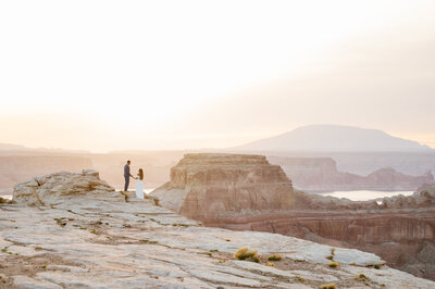 Eloping couple walks hand in hand away from an alpine lake with mountains in the background.