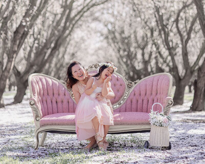 Mom and daughter laughing together on pink sofa placed in a beautiful blooming almond orchard during a Bay Area family photoshoot