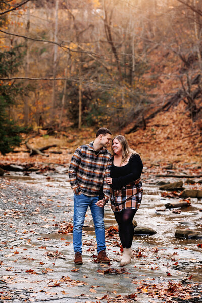 Snuggled up to his arm, a woman gazes at her fiancé surrounded by a creek and fall foliage 