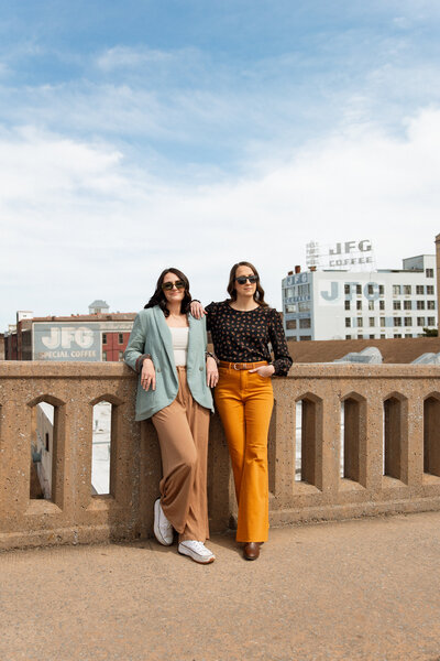 two women leaning against a tan railing with a city background behind them while wearing sunglasses on a sunny day.