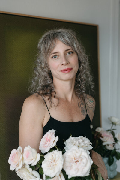 A natural light portrait of a female florist with curly gray hair holding soft blush and white garden roses, photographed indoors against a muted green backdrop. She wears a simple black top and looks warmly at the camera, creating an elegant, editorial-style brand image that highlights fine-art floral design and romantic wedding roses.