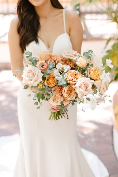 Bride holding a romantic bouquet of peach and blush roses, accented with eucalyptus greenery and delicate white blooms, perfect for an elegant wedding look.