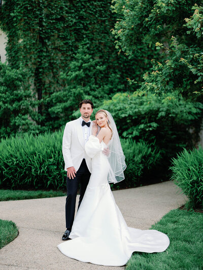 Bride and Groom standing next to each other. Groom is wearing a white tux jacket with a black bowtie  and black trousers and the bride is wearing a dress with puff sleeves and her veil is satin trimmed.