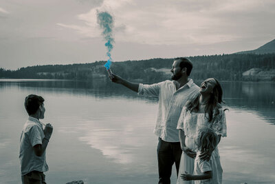 Family celebrating a gender reveal with blue smoke by a serene lake, captured by Joseph Mayden during an emotional and joyful outdoor family photo session.