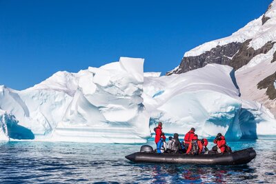 A group of people wearing red jackets are in a black inflatable boat on calm blue water, observing large, white icebergs and snow-covered cliffs under a clear blue sky.