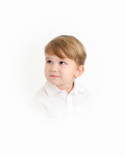 A little boy wearing a crisp white shirt looks to the side in a clean and timeless studio portrait.