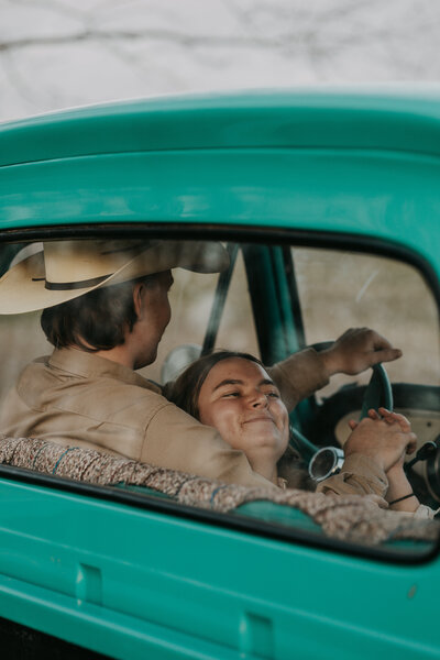 Couple in vintage truck, holding hands and smiling. Romantic, country-style moment with a cowboy hat and rustic charm.