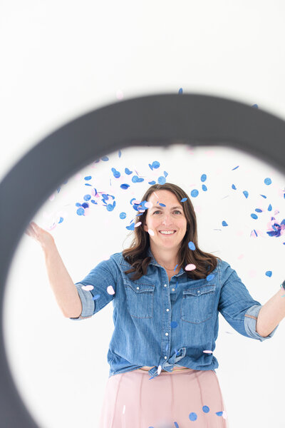 course creator and social media strategist stands in front of a ring of light and is playing with blue and pink confetti. she is holding her hands up and is dressed in a blue jean shirt