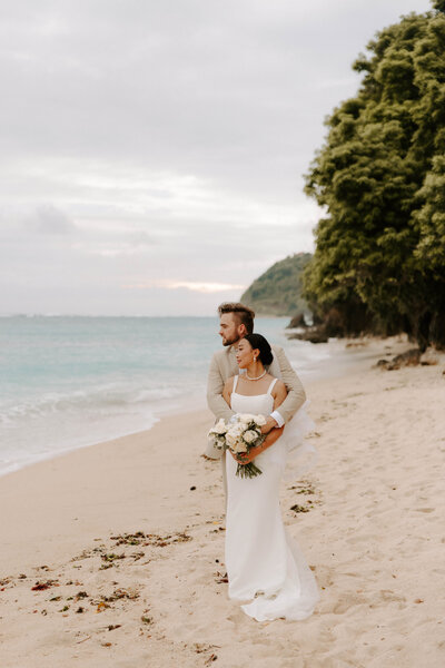 Groom, wraps his arms around his bride who is holding her wedding bouquet, on a beach in Bali Indonesia 