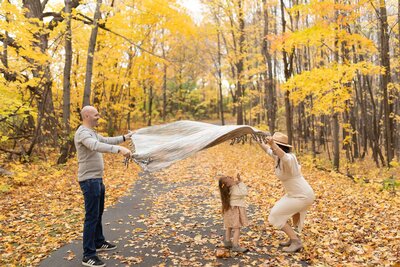 Playful outdoor family session in Maple Grove, Minnesota with parents lifting a blanket while their toddler runs through autumn leaves.