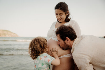 man and child kissing a woman's pregnant stomach on the beach