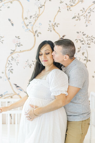 A couple stands in a pink nursery in front of a white crib. The husband stands behind his pregnant wife while wrapping his arms around her belly and kissing her on the cheek. The mother, dressed in a long white dress, closes her eyes, in this sweet shot captured by a Leander Maternity Photographer.