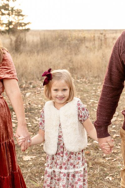 young girl holding parents hands and smiling up at camera during fall family photo session.