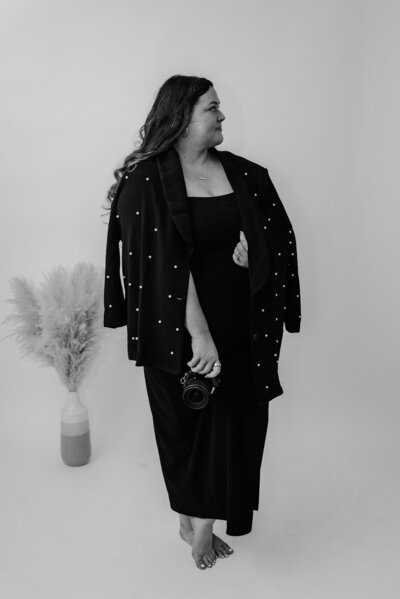 Black and white portrait of an individual wearing a pearl-embellished black jacket and a long black dress, posing confidently while holding a camera with modern pampas grass decor—capturing a bold and stylish fashion-forward aesthetic perfect for branding photography. 