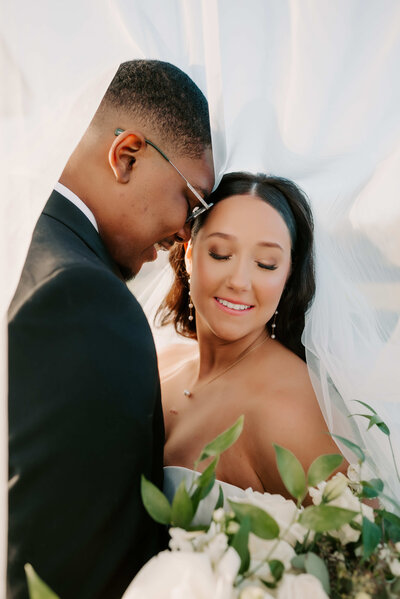 Romantic wedding portrait of a bride and groom embracing under the bride’s veil, with the bride smiling softly while holding a bouquet of white flowers and greenery.