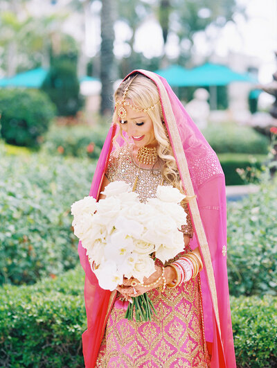 Bride dressed in a vibrant pink and gold lehenga holding a bouquet of white roses before her Indian wedding ceremony at the Ritz Carlton Sarasota — fine-art portrait captured by Asia Pimentel Photography.
