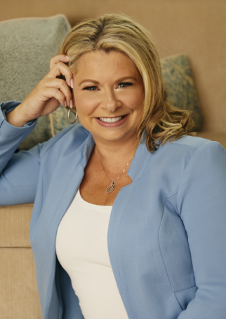 Chanda Bell in a blue suit jacket and white shirt. She is leaning up against a tan sofa with her hand on her temple. She is smiling.