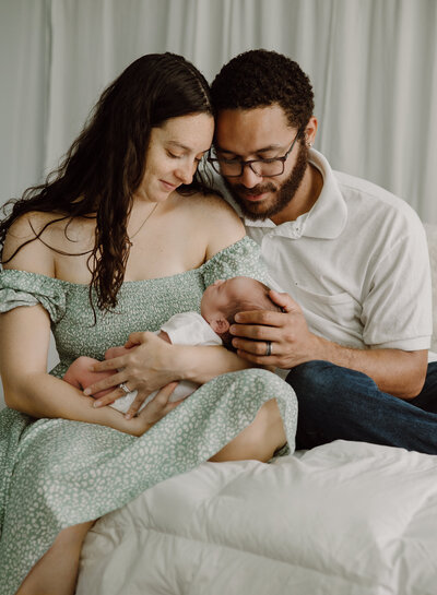 Mom in green dress holding newborn baby boy with husband sitting next to her on a bed in a lifestyle studio in Annapolis Maryland.