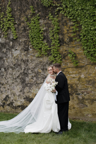 A romantic, cinematic wedding portrait of a bride and groom captured by a luxury wedding photographer. The bride, holding an elegant bouquet, gazes softly away as the groom gently kisses her cheek, set against a rustic, ivy-covered stone wall.