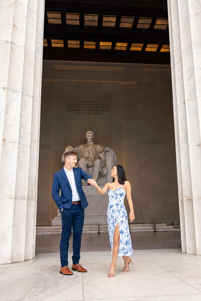 A man in a blue suit and a woman in a floral dress hold hands and smile at each other in front of the Lincoln Memorial statue in Washington, D.C.