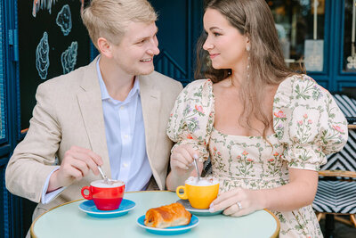 Couple holding coffee cups and gazing at each other inside a colorful Parisian café near Luxembourg Gardens in the Latin Quarter during a romantic photoshoot.