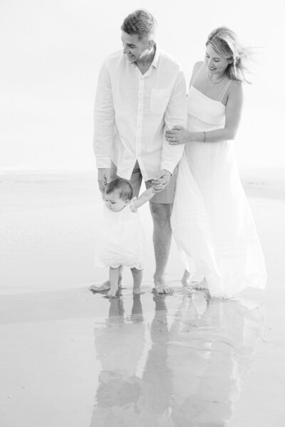 A family walks barefoot on the beach, holding their toddler's hands