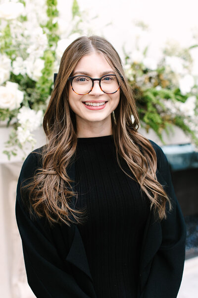 Sydney Russo, senior event sales coordinator at SoHo63, a Chandler wedding and event venue, smiles in front of a lush floral arrangement. Her warm, professional presence represents the expert planning and personalized service SoHo63 offers for weddings and events.