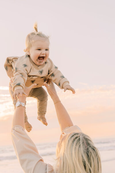 Laughing baby in heart-print outfit lifted in the air during a joyful morning at the beach.
