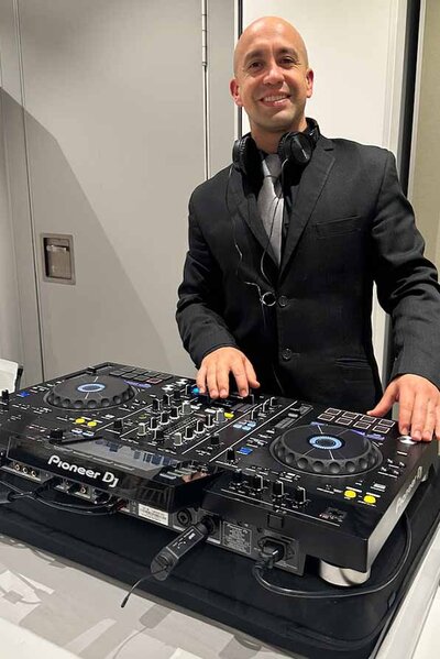Male DJ in a black suit smiling behind a Pioneer DJ controller at a formal indoor event.