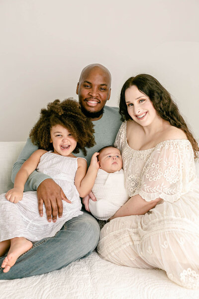 Mom, Dad, Baby and Toddler snuggling on a bed together at their newborn photo session.  Mom is in a beautiful cream lace vici gown, baby is wrapped in white, toddler girl is in a white dress and dad is wearing a teal sweater and light blue jeans. They are smiling and looking at the camera. Baby's eyes are open and he is looking up towards mom.