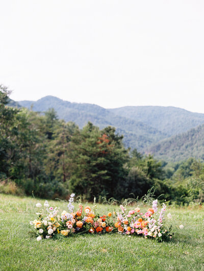 The ceremony arch, or a semi-circle arrangement of beautiful flowers sits in the grassy field with the hills of North Carolina in the background, at wedding venue Paint Rock Farm, on medium format film by Megan Lynn of My Sun and Stars Co.