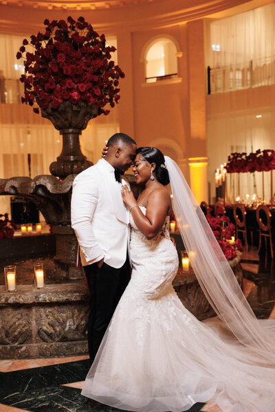 A wedding bride and groom smiling while standing and holding each other with their foreheads touching.