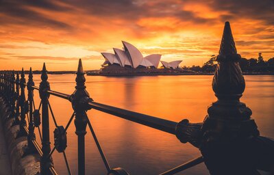 A breathtaking view of the Sydney Opera House at sunset, with its white sails glowing against the orange and golden hues of the sky. The serene water and intricate railing add to the beauty of this unforgettable destination, showcasing the global adventures made possible through vacation fundraising for charities.