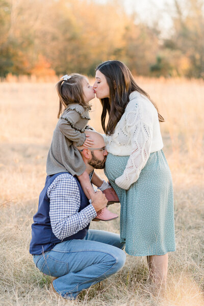 Outdoor field maternity session in the fall with mom, dad, and toddler girl. Toddler girl is on dad's shoulders while mom kisses toddler and dad kisses mom's belly. Brandon, MS Maternity Photographer, CJ and Olive Photography.