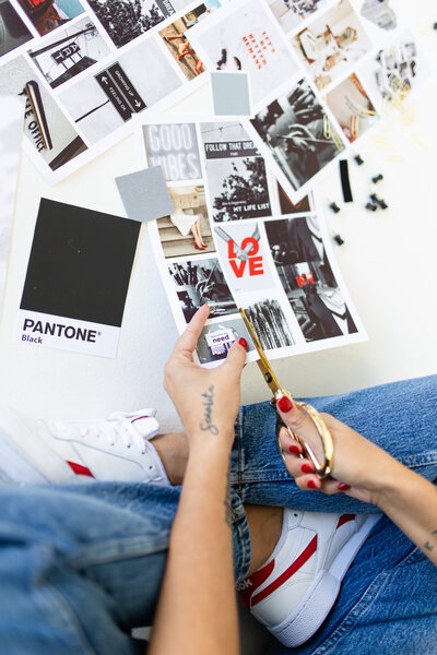 A woman is cutting with gold scissors a print paper with brand moodboard images. Next to her hand is a paper with black pantone.