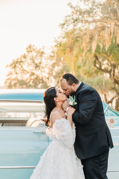 Bride and groom kissing in front of a vintage car during a sunset, in love