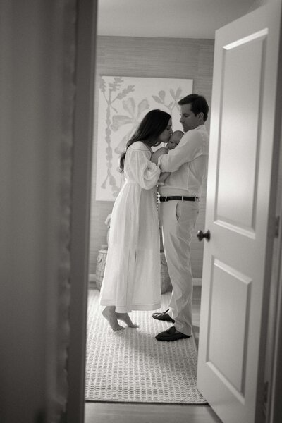 Black and white photo through a doorframe of father holding newborn while mom kisses the baby