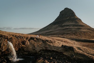 Bride and groom hold hands while walking around a grassy hill during their elopement in Iceland. 
