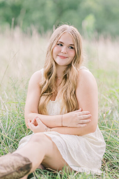 A high school senior sitting in tall grass, smiling softly in a white dress and boots during golden hour — Raleigh portrait photos.