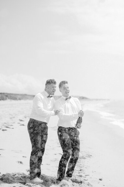 Two grooms celebrating on the beach in Cape Cod, laughing and enjoying their wedding day. Captured by Felisha Lees Photography, this moment embodies joy, connection, and love.