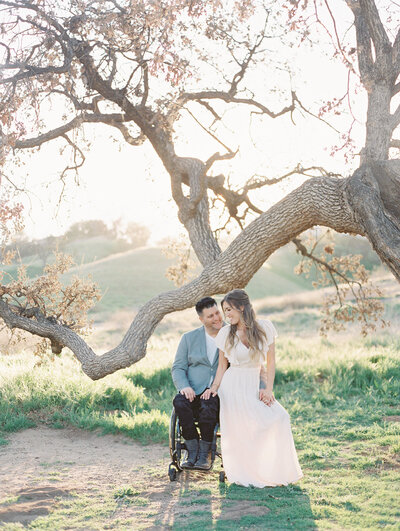 Photographer, Tiffany Gentry, sits on her husband's lap in his wheelchair under beautiful oak tree