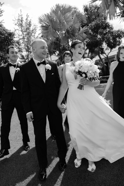 Bride laughing while holding her bouquet, walking hand in hand with the groom and surrounded by the wedding party.