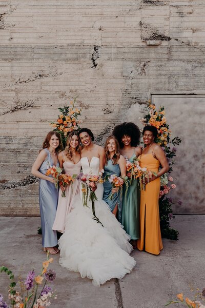 Bride and bridesmaids casually hug while wearing different colored dresses