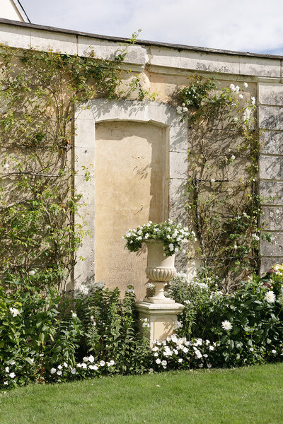 A stone wall with climbing vines and a floral urn sits in a tranquil courtyard