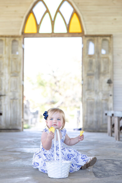 flower girl at wedding sits in aisle at Gruene Estates in New Braunfels with blue floral dress and yellow petals in hands