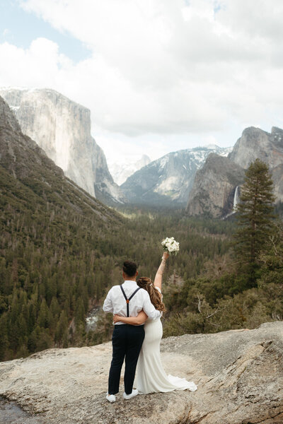Bride and groom stand at their Yosemite Wedding Location: Tunnel View and bride holds her bouquet up in celebration