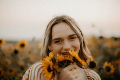 Photo of a senior girl holding a sunflower up to her face and smiling at the camera.
