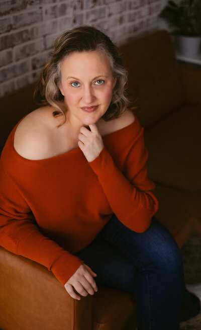 Bentonville boudoir photographer Wildflower Boudoir wears an orange sweater while posing on a couch .