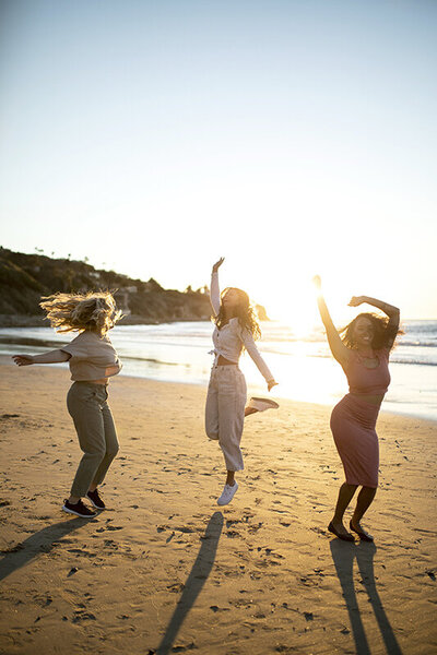 Trois femmes dansant sur la plage joyeusement, bras levés.