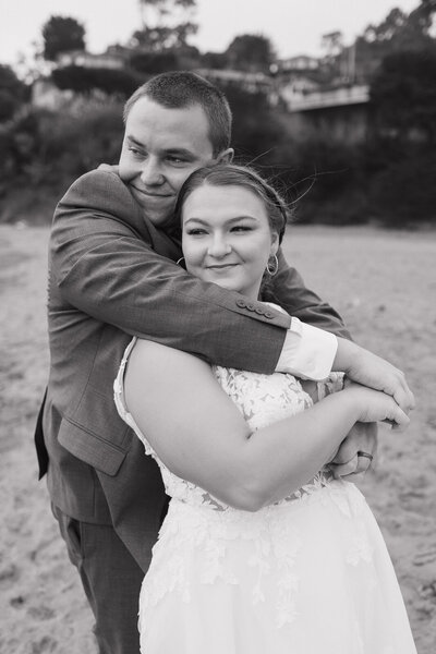 black and white photo of a couple on the beach for a wedding, his arms draped around hers and soft smiles